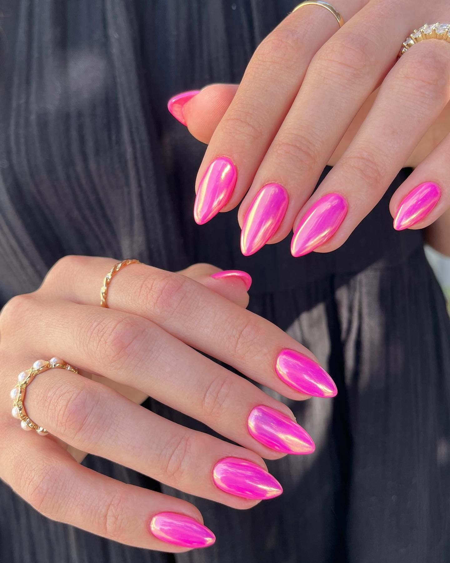 Long pointed almond nails in iridescent fuchsia chrome polish, paired with delicate gold rings on fair hands, set against a dark, softly textured fabric background.

