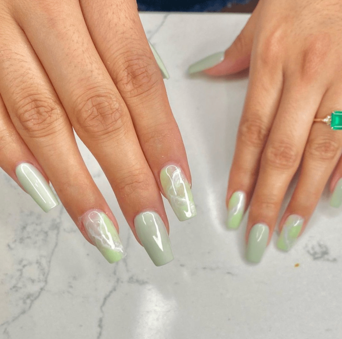 Two hands with pastel green glass sage nails featuring white marble accents and a square-cut emerald ring on one finger, set against a marble countertop