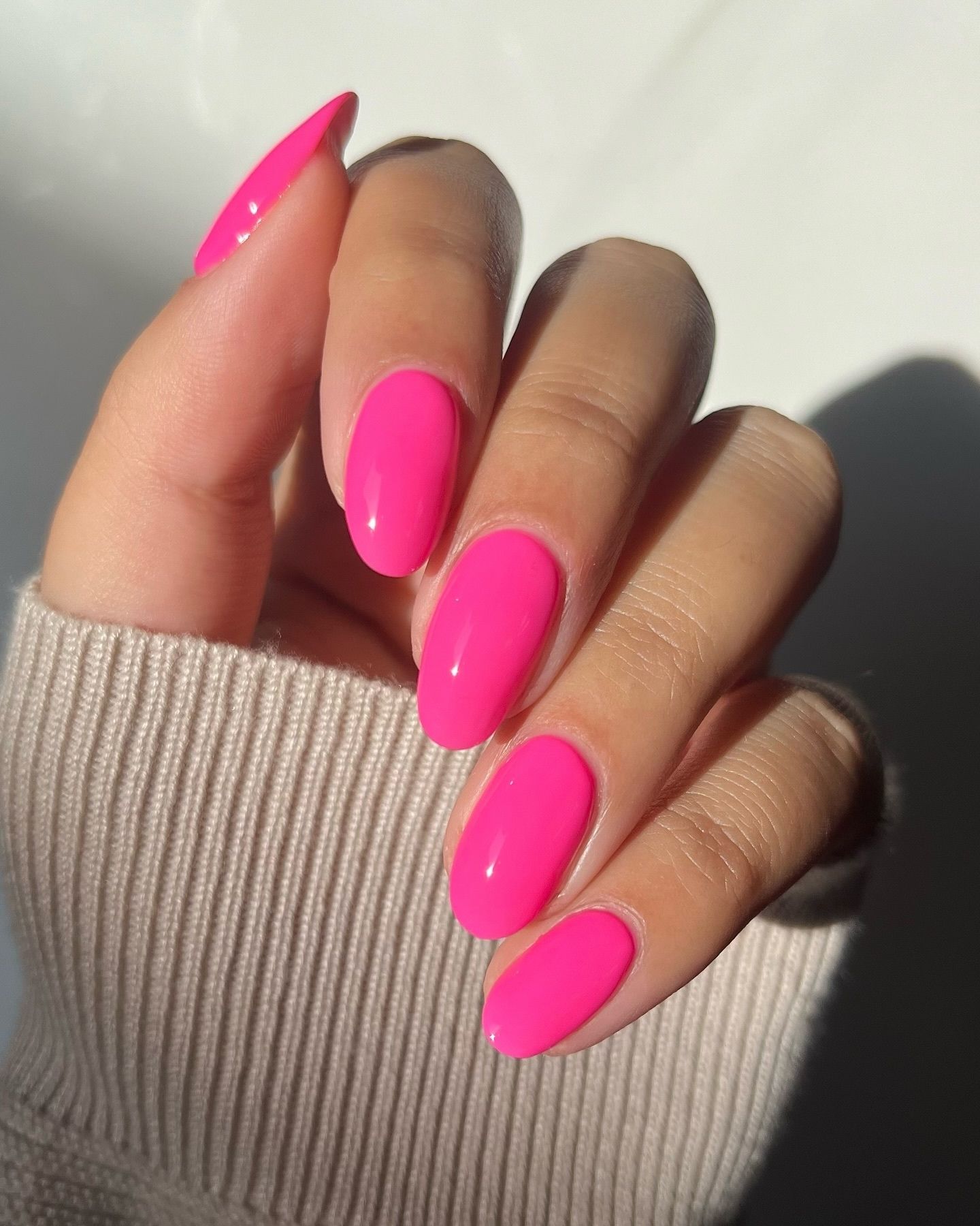 Bright hot pink glossy nails in an oval shape, resting on a light beige knit fabric with soft natural lighting and a clean, minimal background.