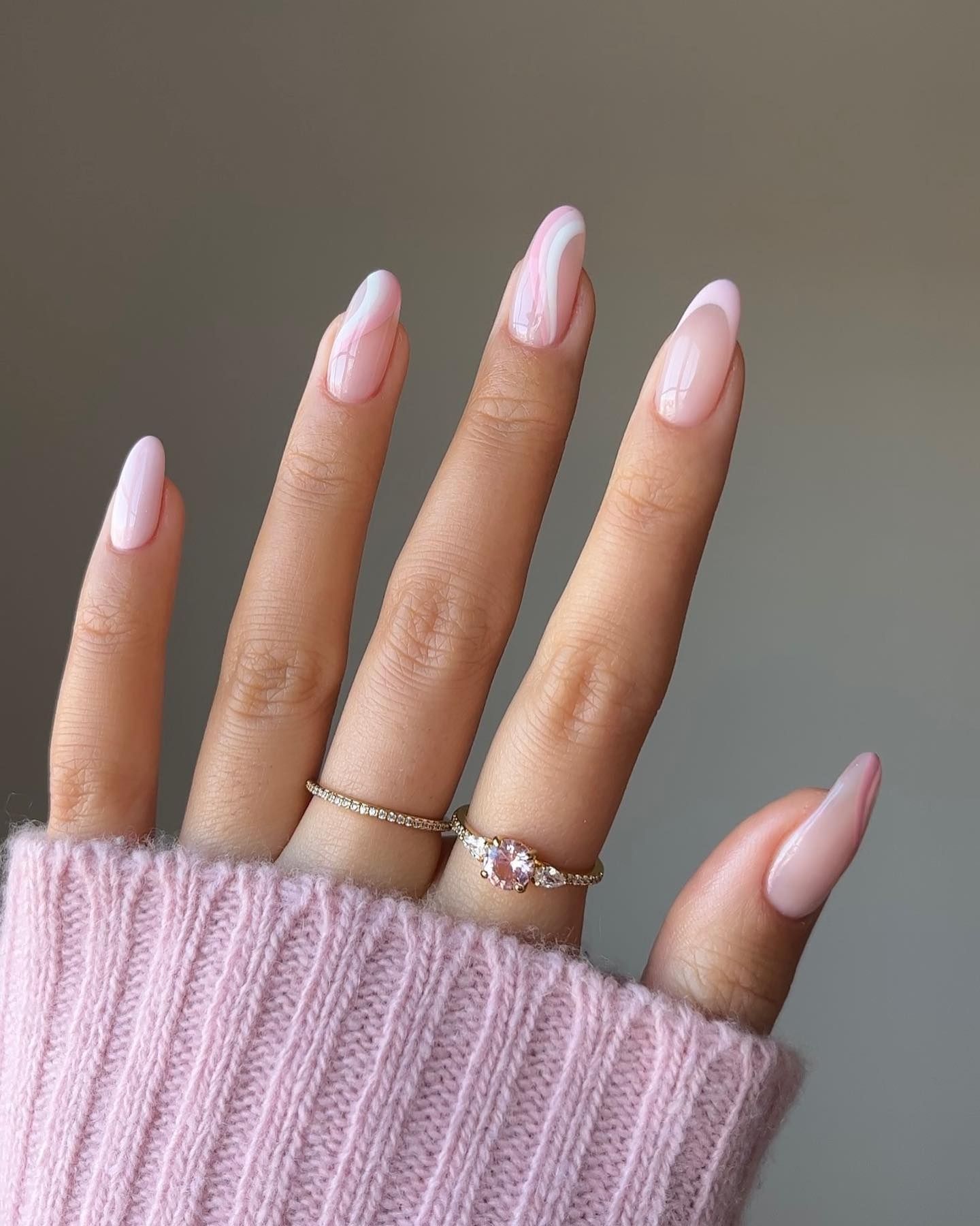 Soft pink glossy nails with elegant white swirl designs on a short almond shape, paired with delicate gemstone rings and a blush pink knit sweater in the background.