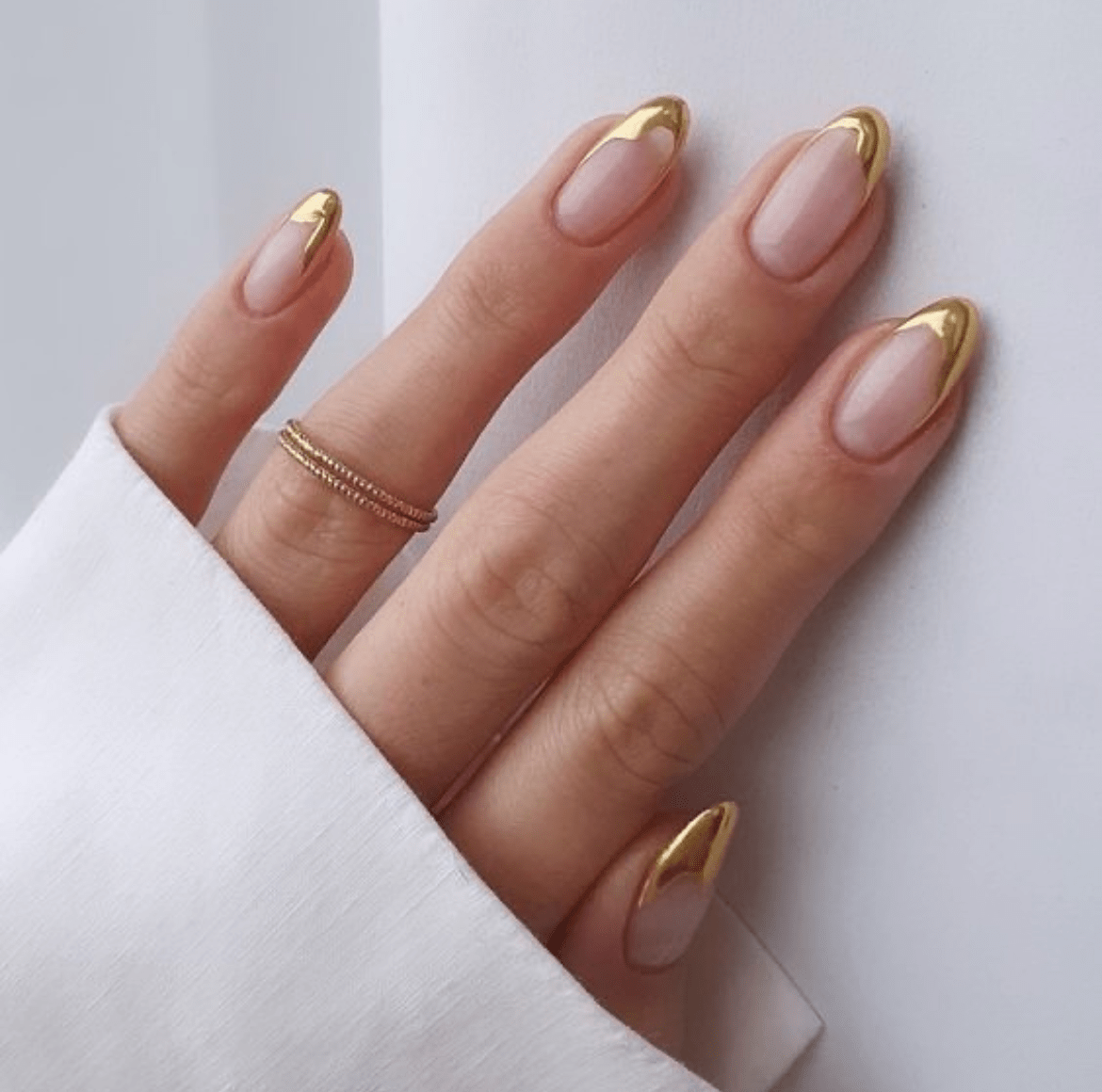 Almond nails with gradient gold metallic tips on a natural pink base, paired with a gold braided ring