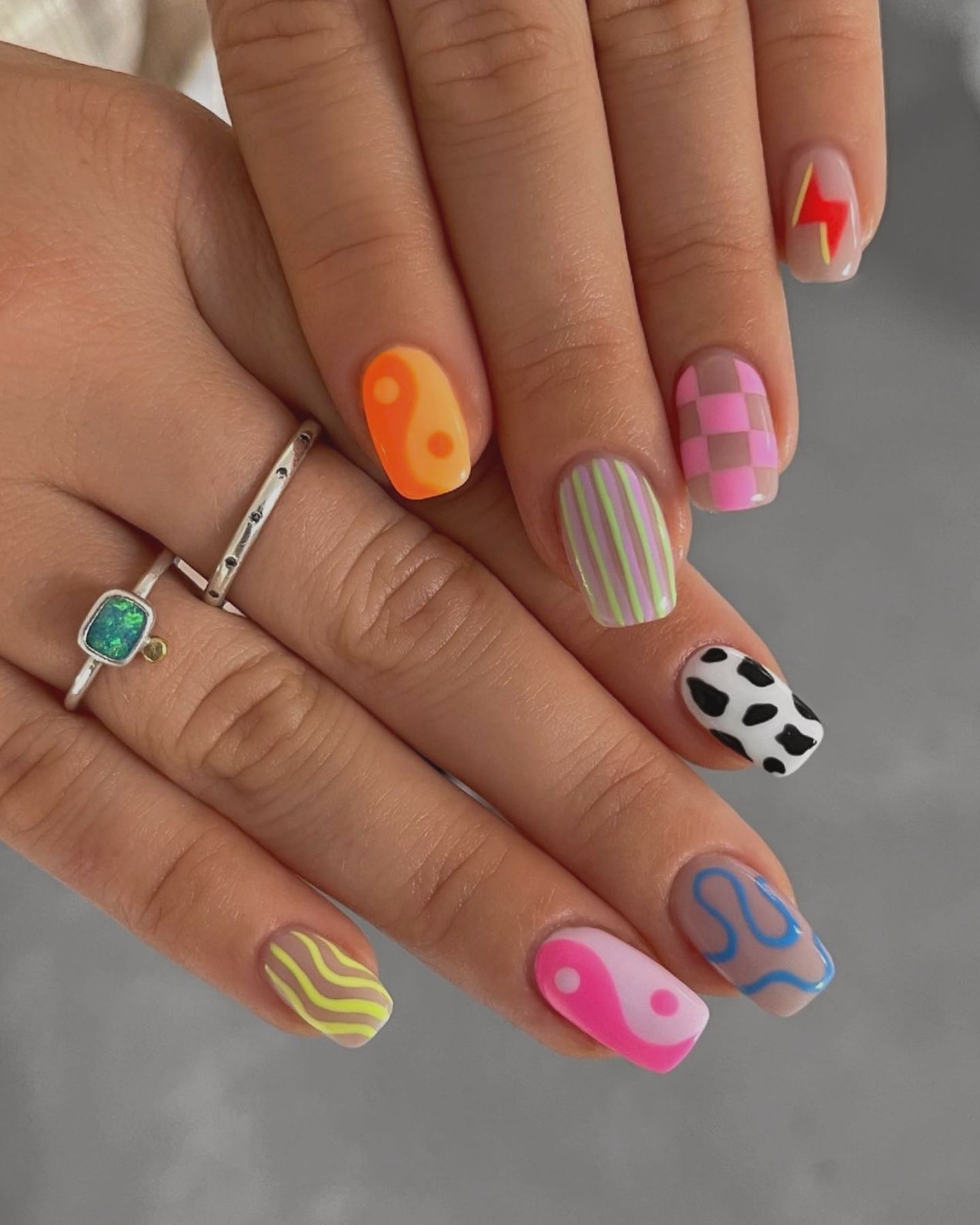 Vibrant summer nails featuring neon green waves, bright pink and orange nails with red accents, pastel pink with blue squiggles, striped pastel nails, black and white cow print, and brownish-purple waves, complemented by a silver ring with green and yellow stones.