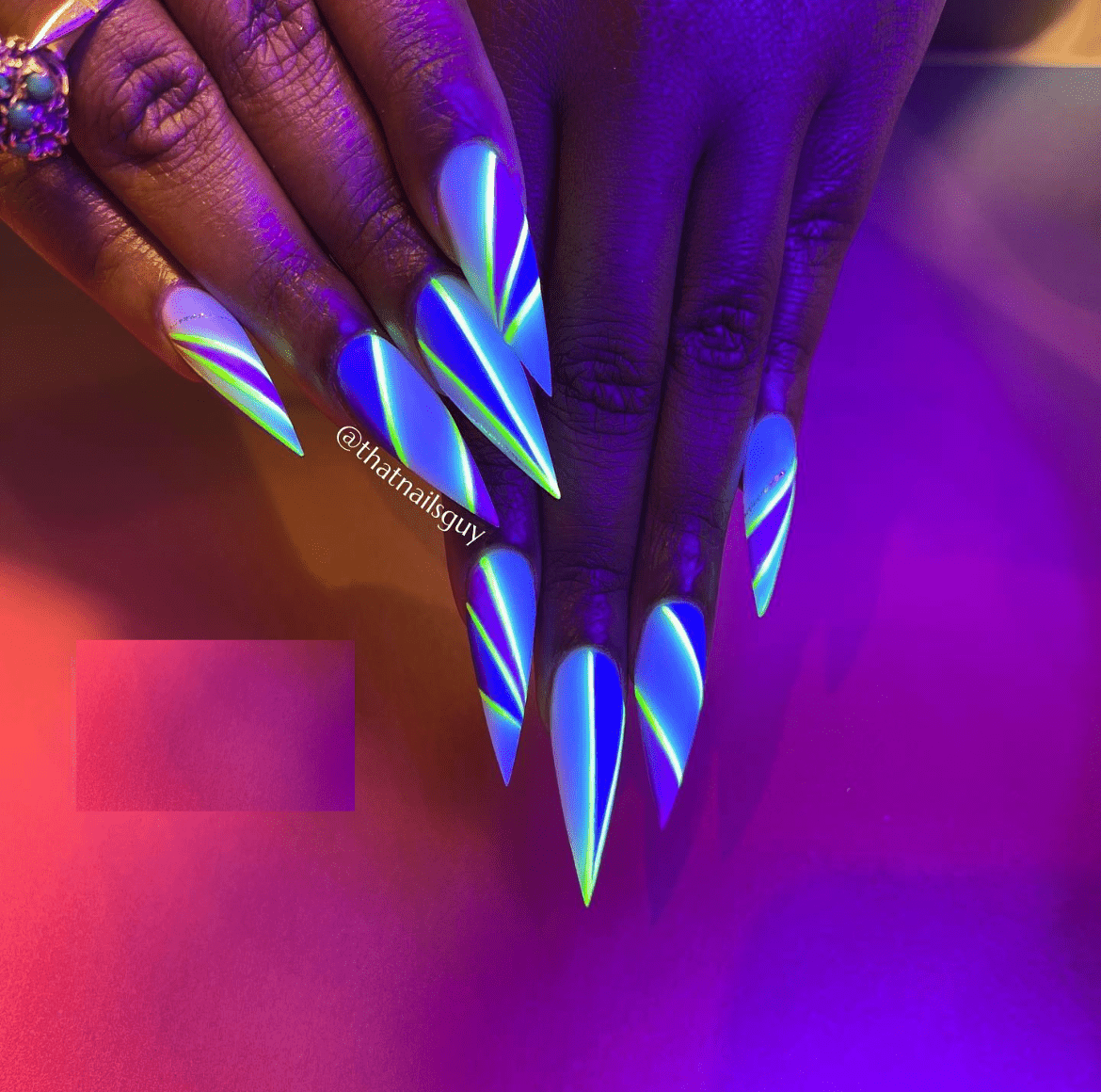 Long pointed nails with glowing neon designs in blue, purple, green, and pink tones, set against a vibrant gradient background with gemstone ring detail