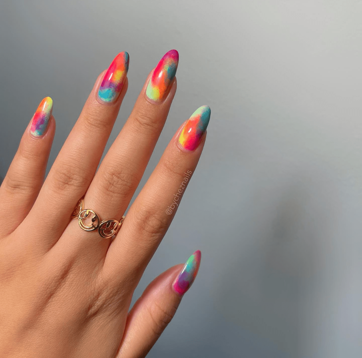 Almond-shaped nails with vibrant watercolor-inspired neon designs in pink, yellow, blue, green, and orange, paired with a stylish gold ring on the middle finger