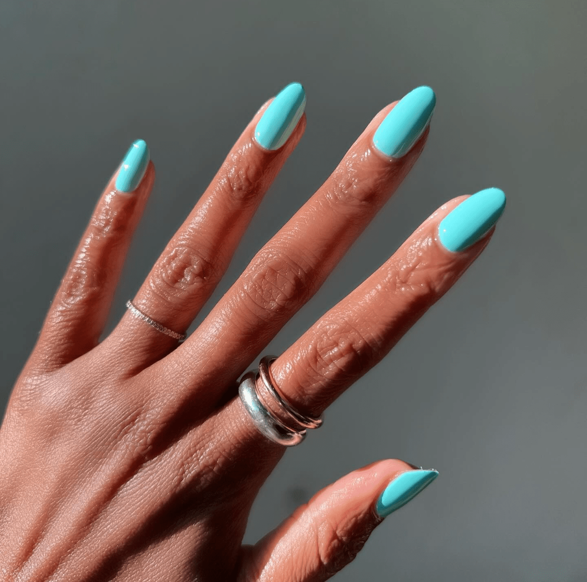 Pastel blue almond-shaped nails with glossy finish, styled with silver and gold rings against a grey background.