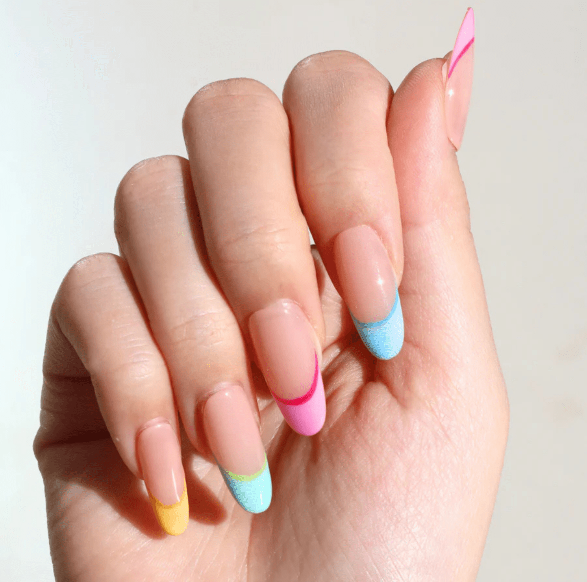 hand with stiletto-shaped nails featuring pastel French tips in pink, yellow, and light blue over a clear base, shown against a light neutral background with the pinky slightly curled