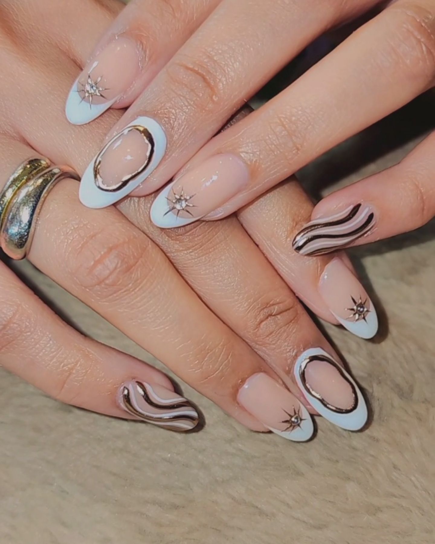 Almond-shaped nails with a clear base, glossy finish, gold gradient stripes, dark oval outlines, sparkling star embellishments on French tips, complemented by a silver ring on the finger.