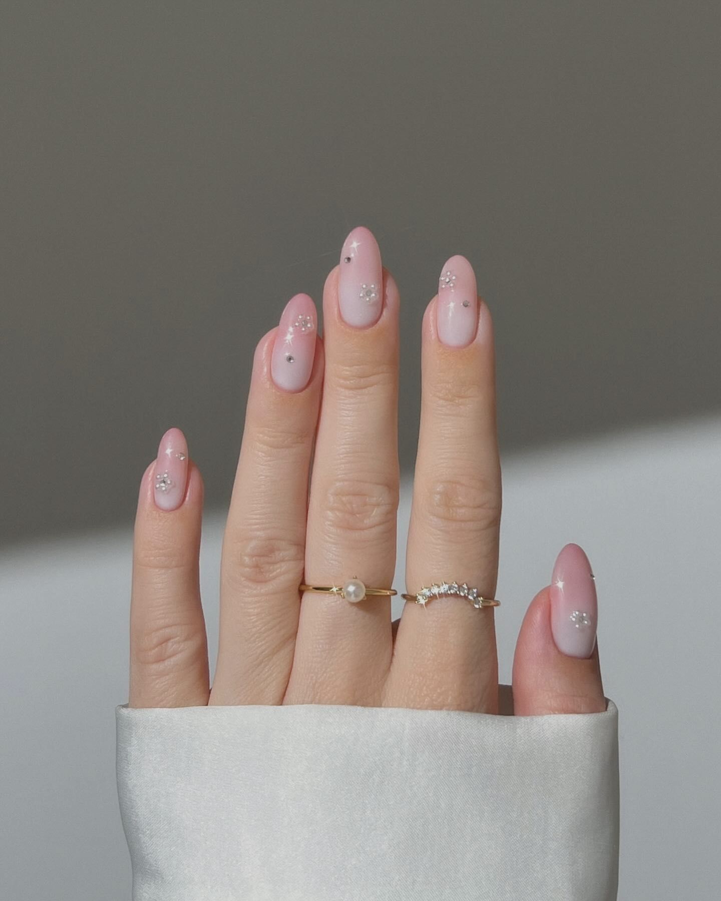 Almond-shaped gradient pink nails with rhinestone accents, styled with gold rings including a pearl and crescent stone band.