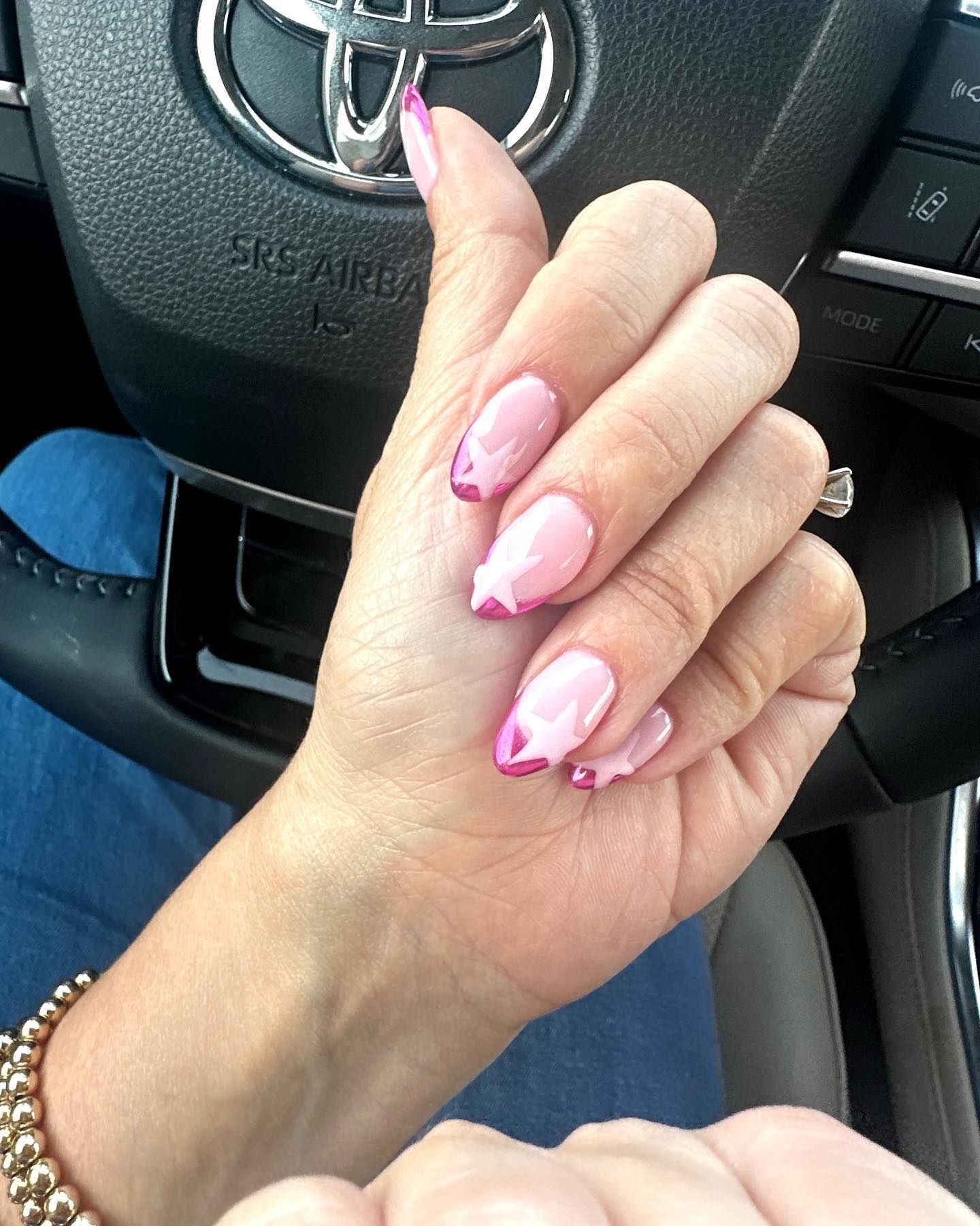 Close-up of pink chrome French tip nails with baby pink stars, worn with a silver ring and gold beaded bracelet, hand resting on a Toyota steering wheel.

