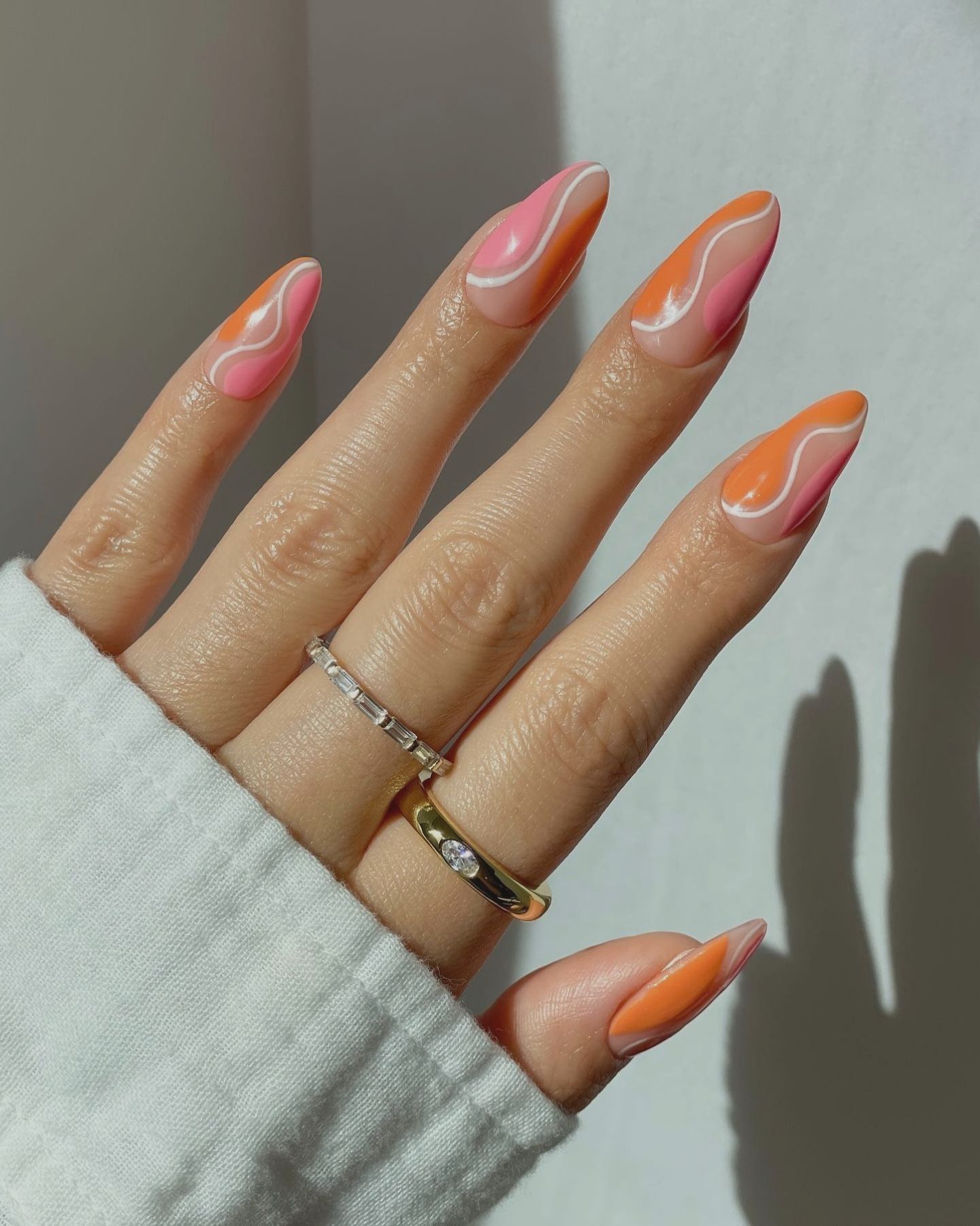 Almond-shaped nails with pink and orange abstract wavy design, styled with diamond rings and a white garment background.