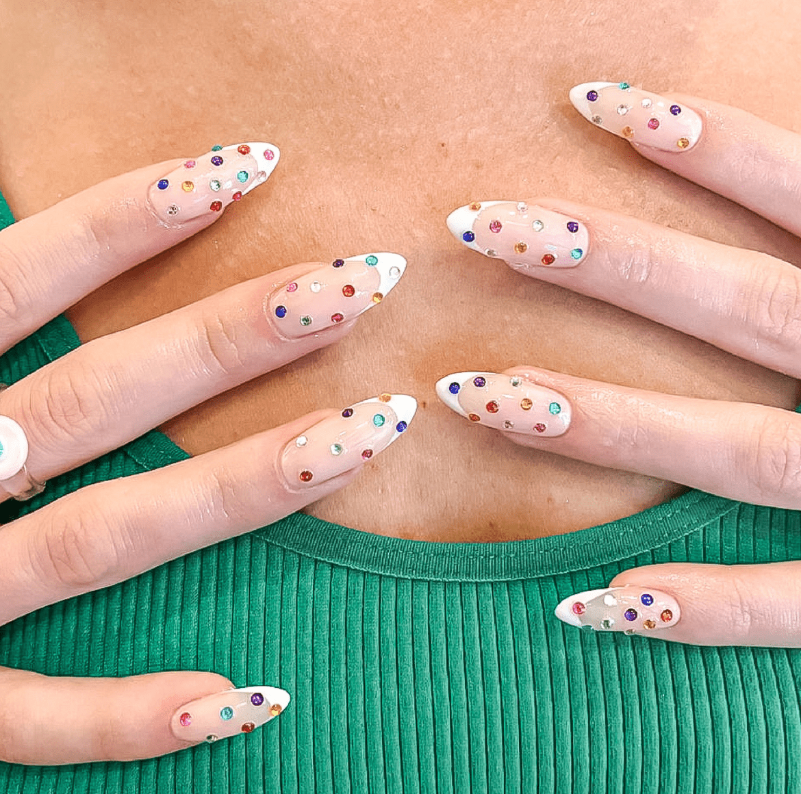 Clear French tip nails decorated with multicolored rhinestones in various shapes and sizes, set against a light pink base with a teal ribbed top in the background.