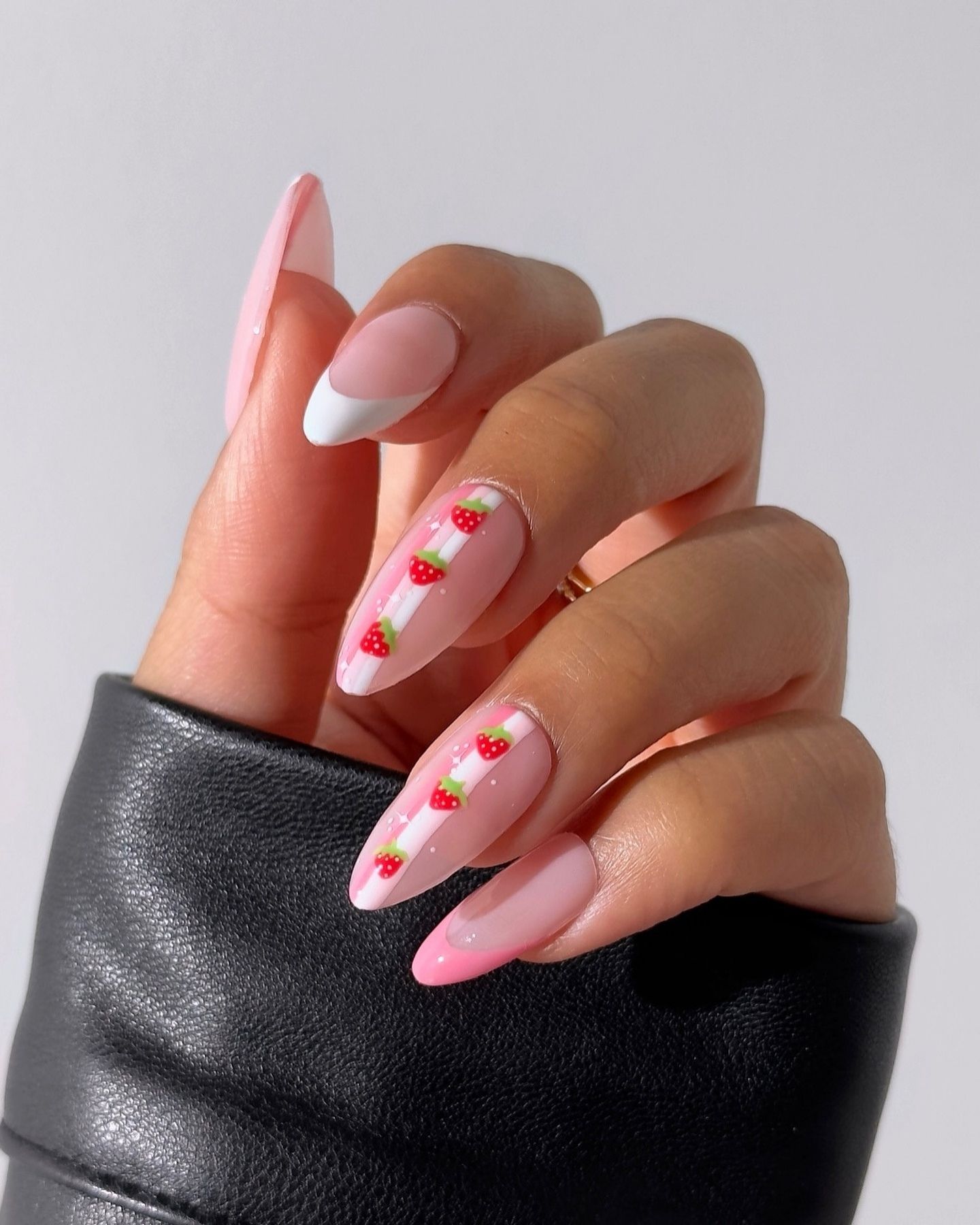Soft pink almond nails with white stripes and small 3D strawberry decorations, accented by a French tip and gold ring, resting on black textured leather.