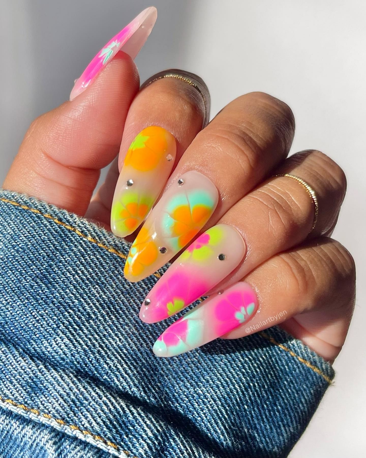 Almond-shaped nails with vibrant tie-dye floral gel design and rhinestone accents, resting on blue denim cuff