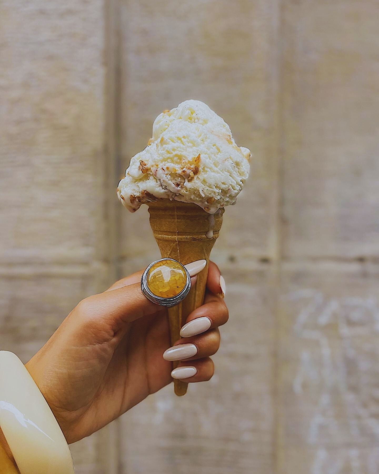 Hand with light-colored vanilla nails holding a waffle cone of white ice cream topped with bits, wearing a silver ring and yellow bangle.