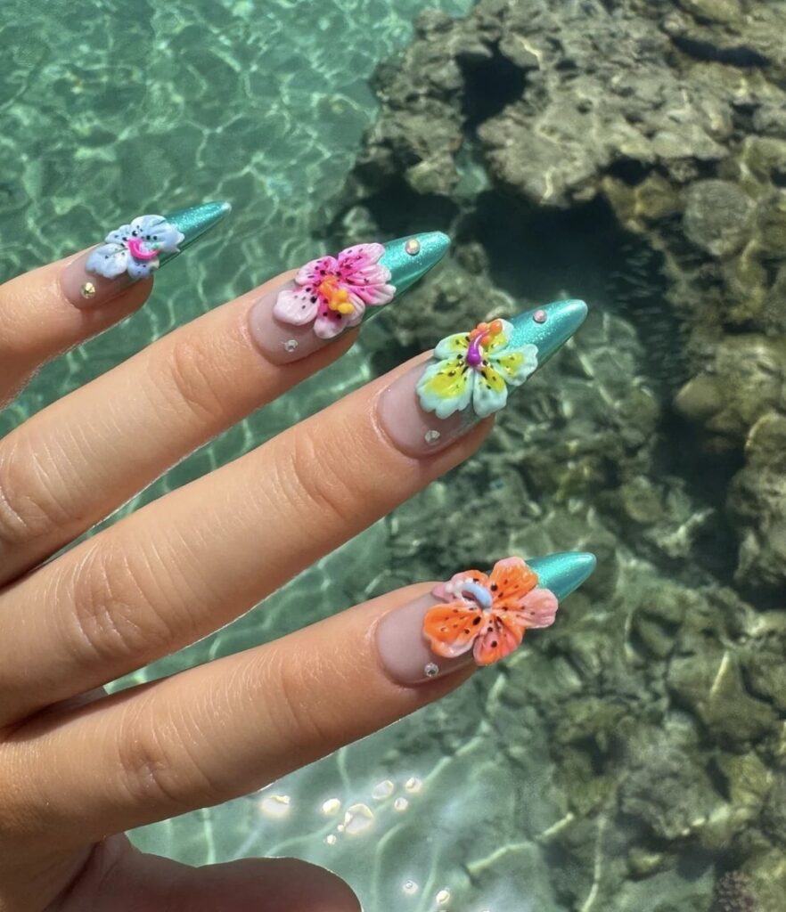 Close-up of long stiletto nails with 3D hand-sculpted hibiscus flowers in tropical colors over a shimmery turquoise base, photographed above clear ocean water.