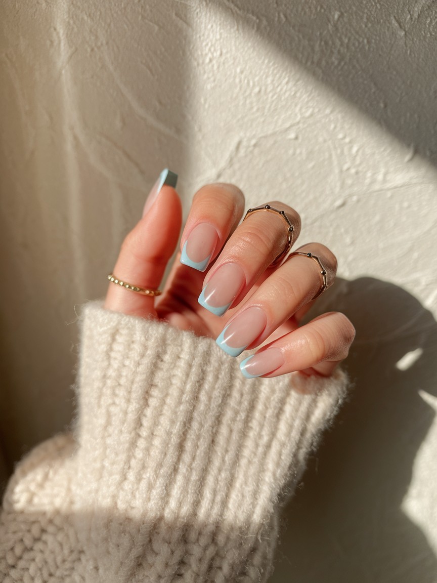 Short nude nails with minimalist baby blue micro French tips, styled with gold rings and a soft knit sleeve.

