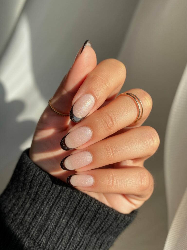 Hand with almond-shaped nails featuring black shimmer French tips, styled with gold rings and a black knit sleeve on a neutral fabric background.