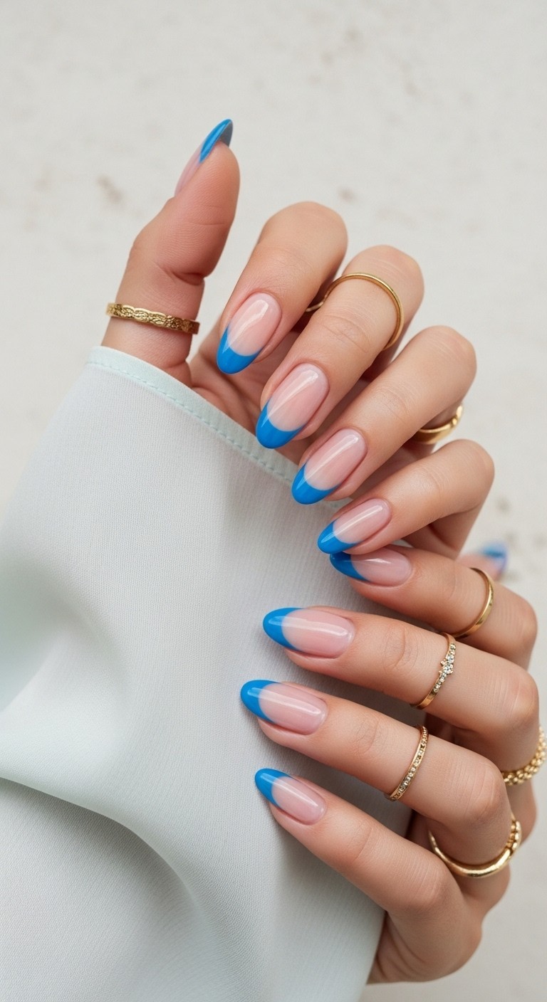 Nails with pastel blue crescent accents at the cuticle line over a sheer nude base, styled with soft lighting and delicate gold rings.

