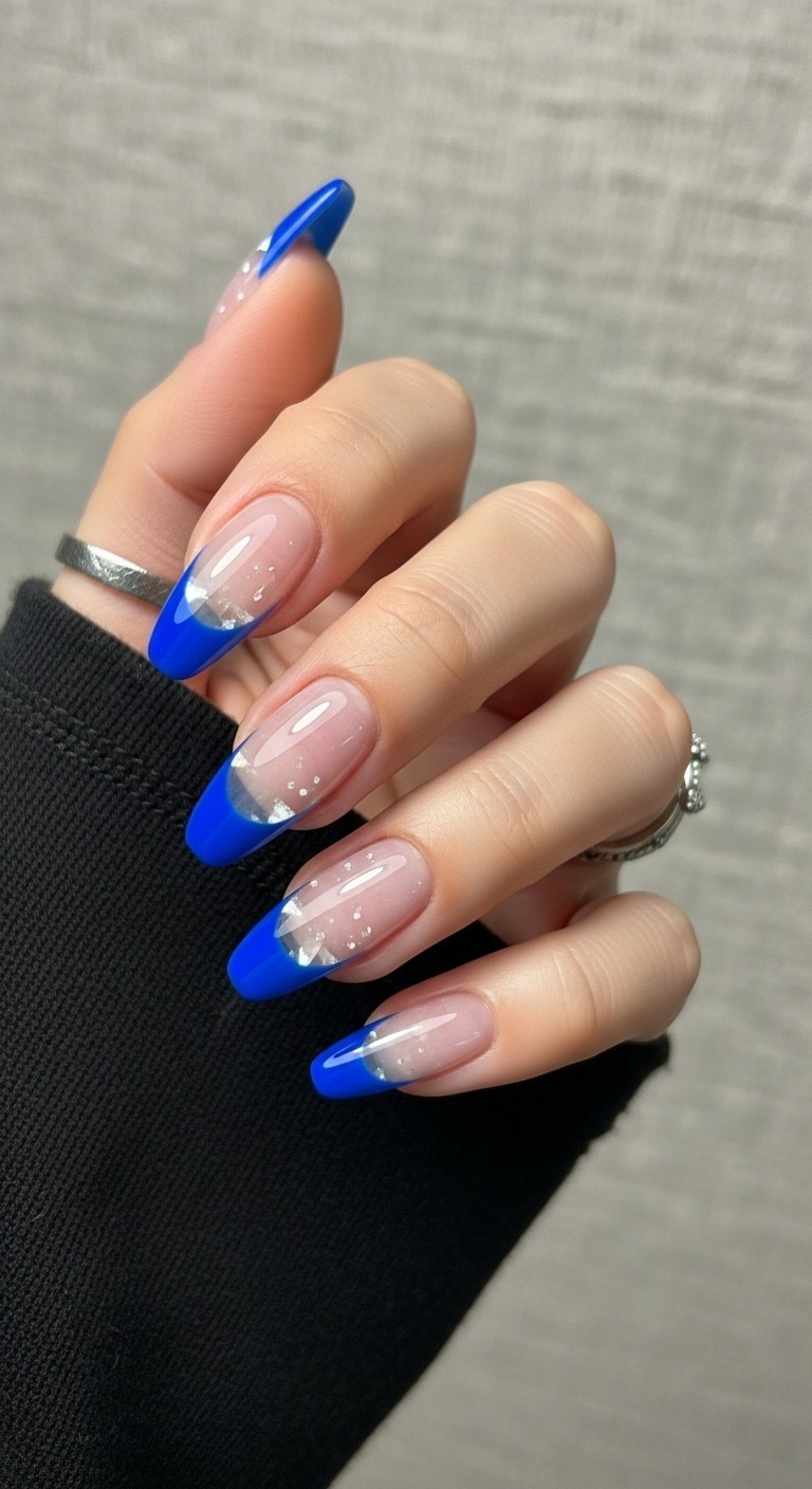 Hand with almond-shaped nails featuring royal blue tips and translucent crystal-clear bases, styled with a light sleeve and neutral background.