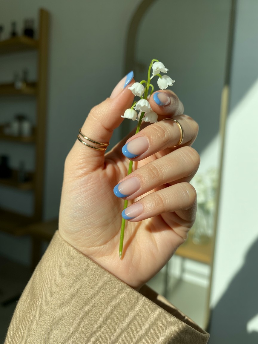 Relaxed hand with medium-length nails featuring cornflower blue smile line French tips on sheer nude base, styled with thin gold rings and linen sleeve against off-white background.