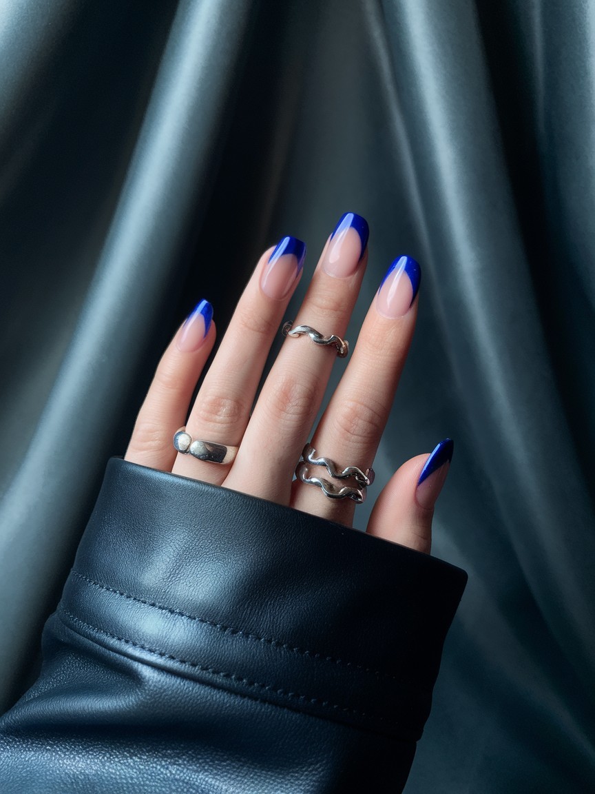 Medium-length nails featuring glossy deep cobalt blue French tips with sheer nude base, accented by silver rings and black leather sleeve.

