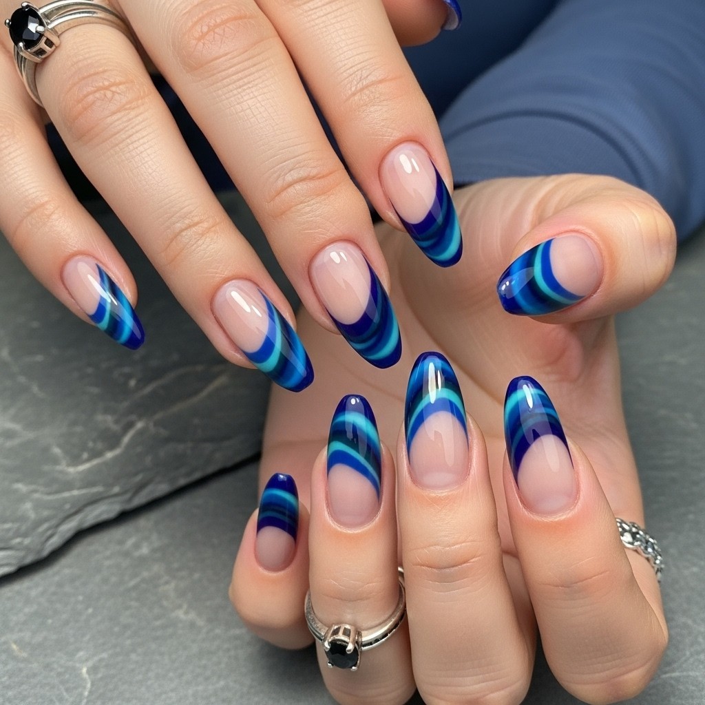 Almond-shaped nails with deep navy and teal swirl French tips on a sheer nude base, styled with silver rings and a cool-toned background.