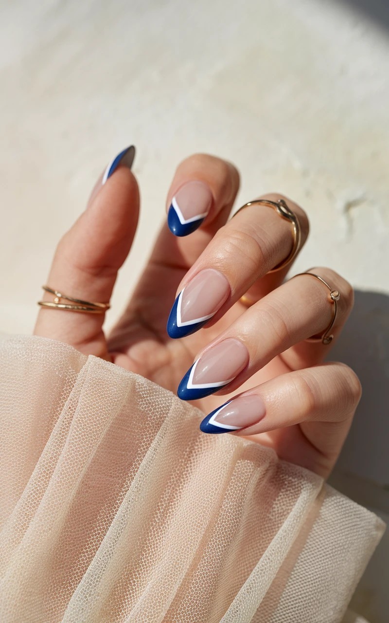 Almond-shaped nails with deep V-shaped cobalt blue French tips on a nude base, styled with soft lighting and minimal accessories.


