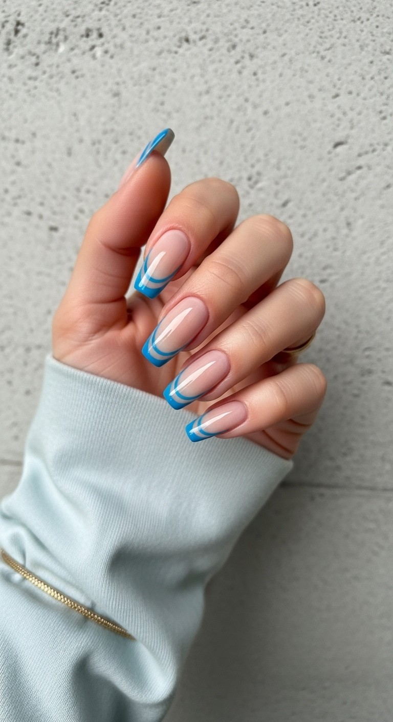 Hand with almond-shaped nails featuring a nude base and double blue French lines, framed by a pastel blue sleeve and soft neutral background.

