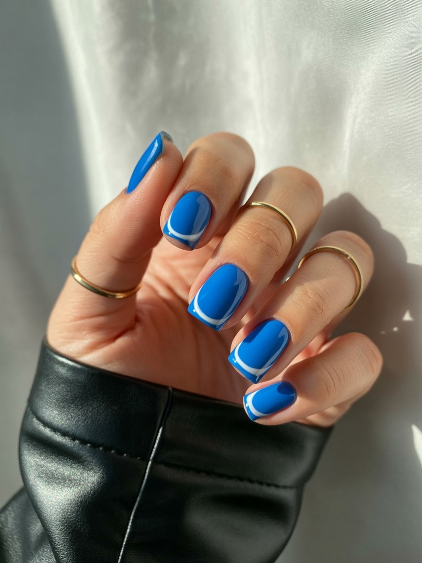 Relaxed hand with medium-length nails featuring vibrant electric blue angular French tips, styled with thin gold rings and black leather sleeve against off-white background.

