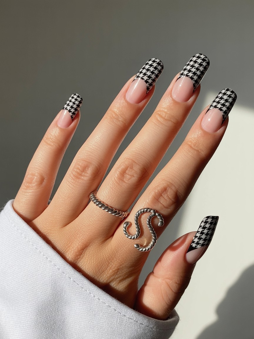 Close-up of long nails featuring black and white houndstooth French design with clear sections and a silver twisted rope-style ring on the middle finger.