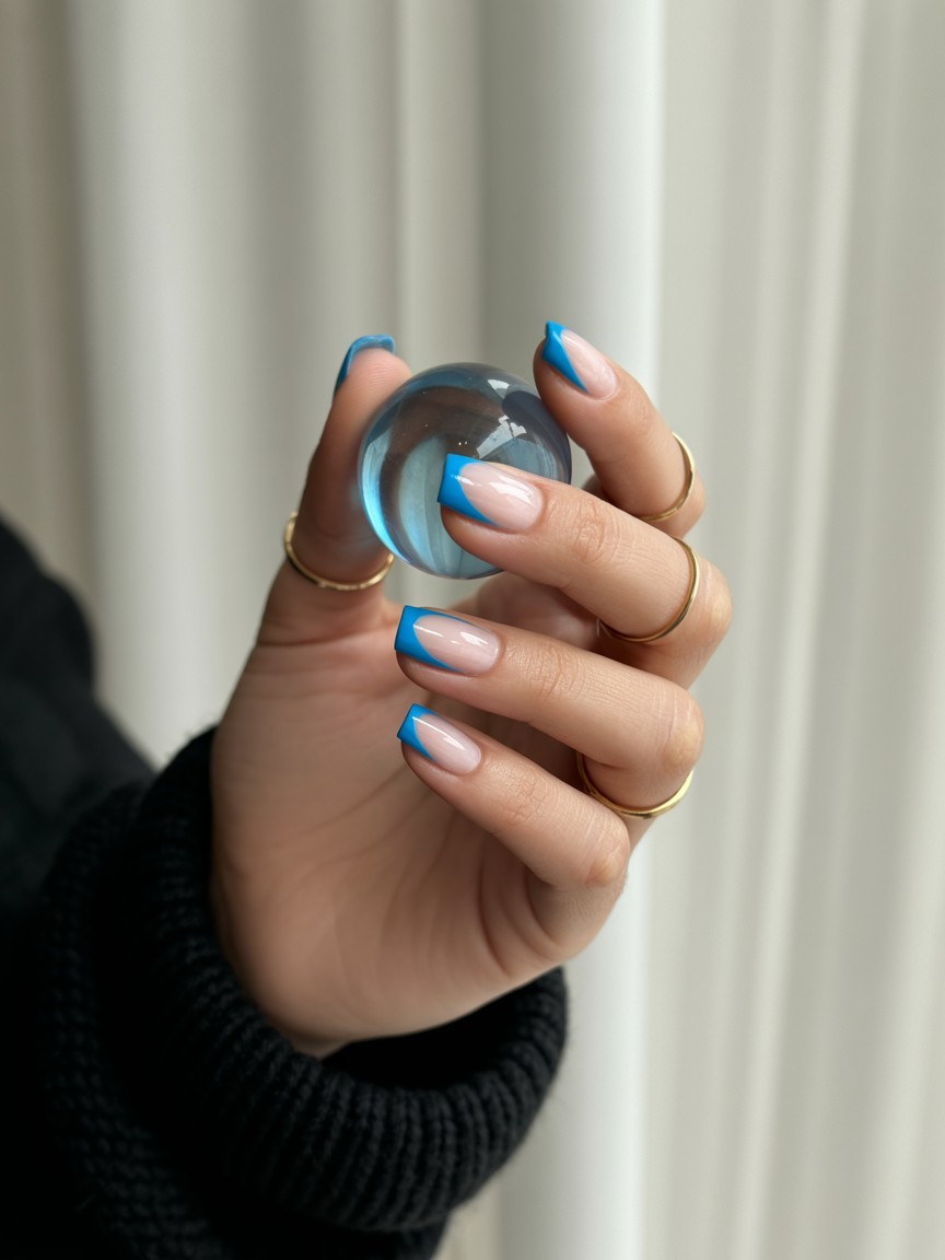Relaxed hand with medium-length nails showcasing bright neon blue French flick tips on sheer nude base, styled with thin gold rings and black knit sleeve against off-white background.

