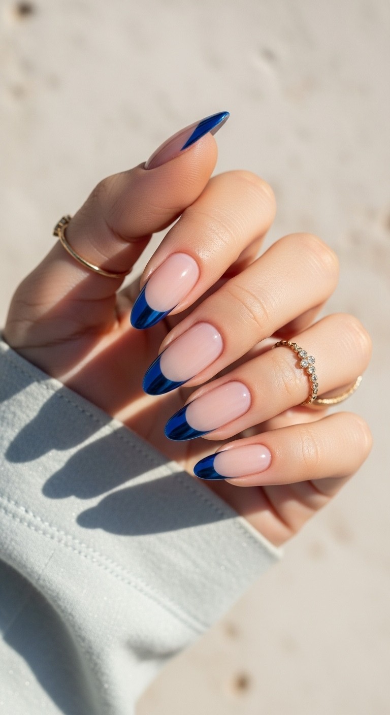 Hand with nude nails featuring bold blue half-moon accents at the base in a reverse French manicure style, styled with soft lighting and minimal accessories.

