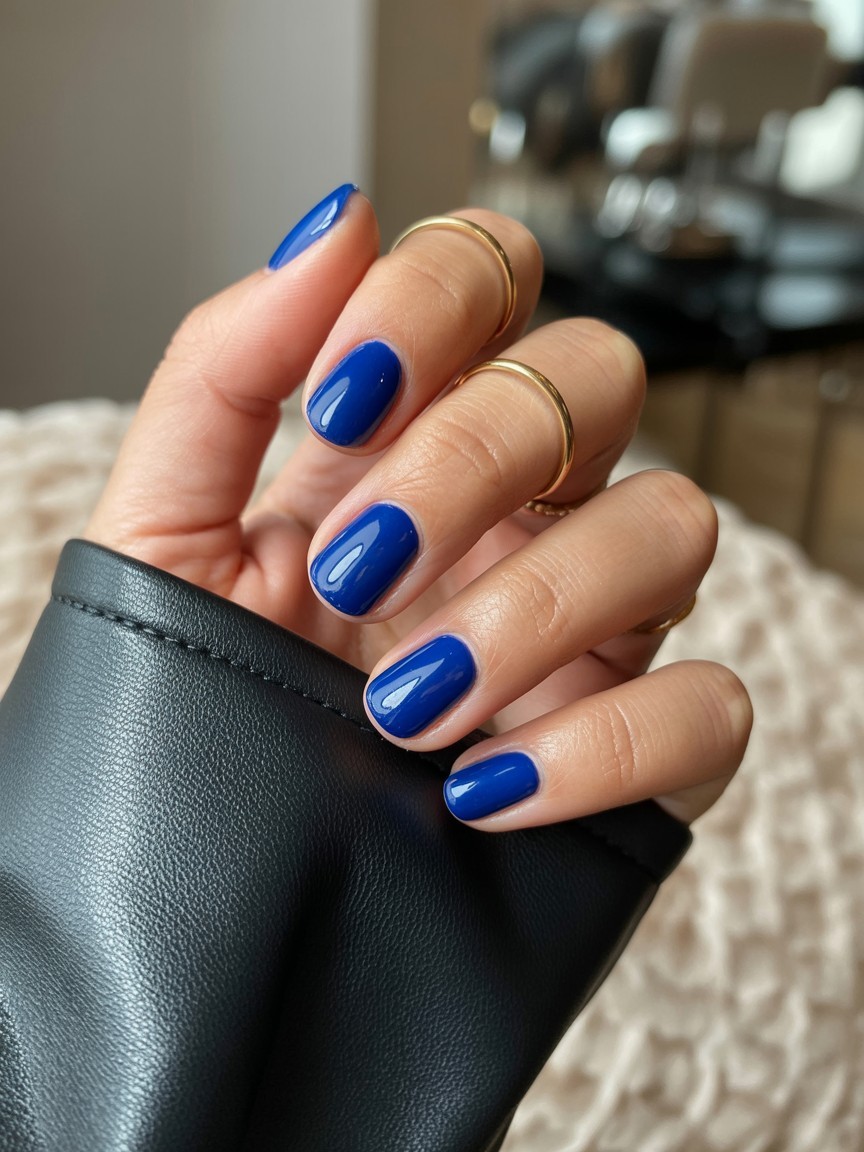 Relaxed hand with medium-length nails featuring glossy royal blue dramatic French tips, styled with minimalist gold rings and black leather sleeve on a neutral textured background.