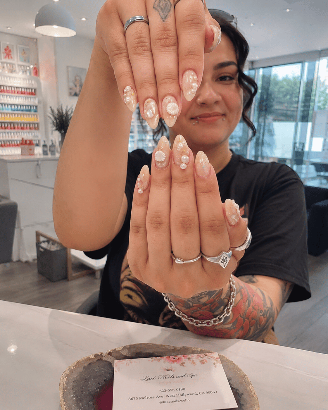 Sandy nude almond-shaped nails with delicate shell art and pearl accents displayed in a bright modern nail salon.