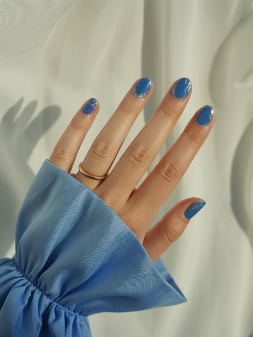Hand with sapphire blue nails featuring foil tip overlays and a glossy finish, complemented by a gold ring and summer dress sleeve.

