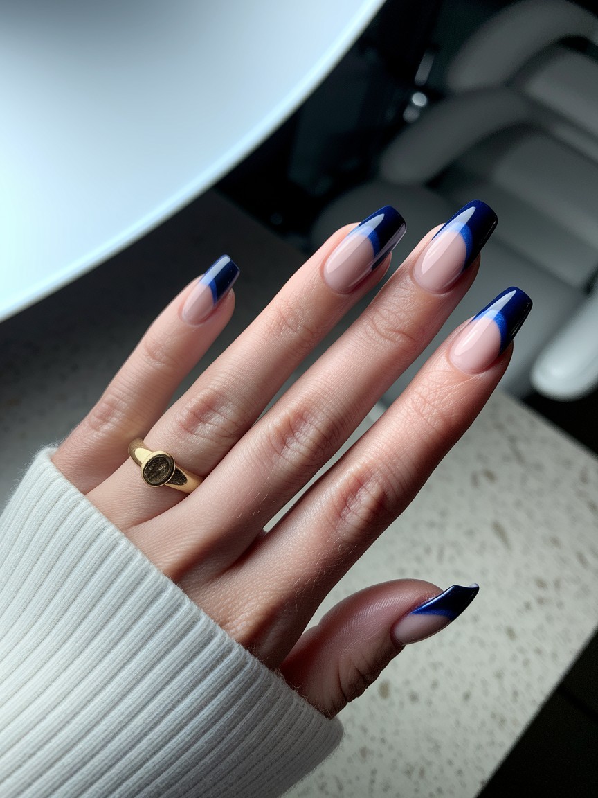 Back of hand with deep sapphire French tip nails, soft lighting and gold ring, resting on stone surface with a blurred salon background.

