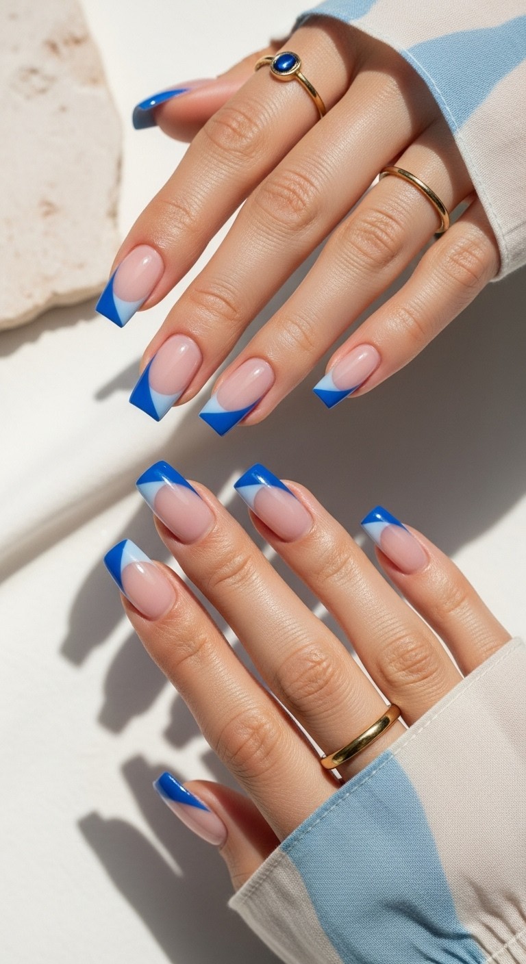 Hand with nude base nails featuring diagonally angled blue French tips, styled with soft lighting and a flowy pastel sleeve.

