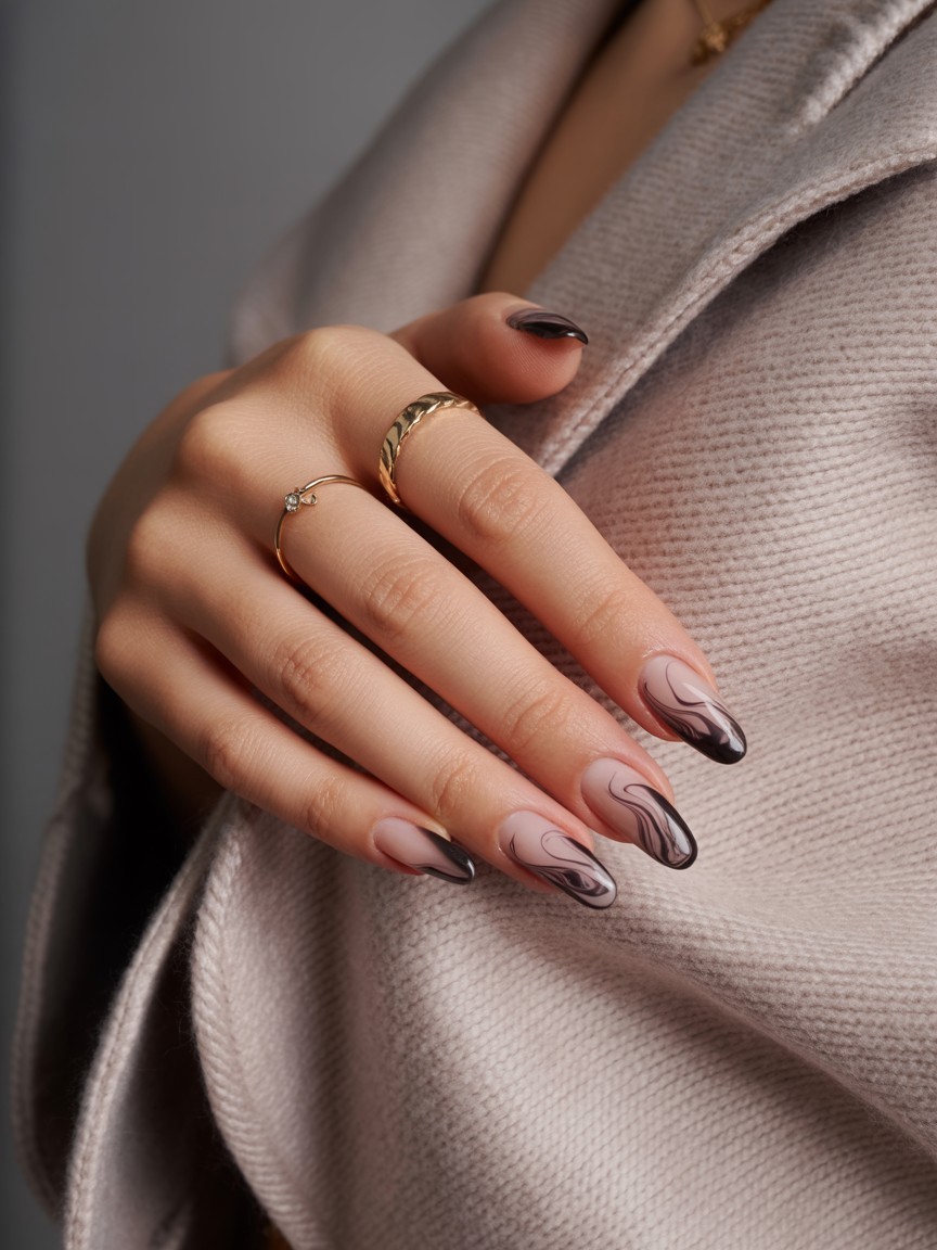 Almond-shaped nails with swirled black French tips resembling ghostly patterns, accented by gold rings and set against a soft grey background.