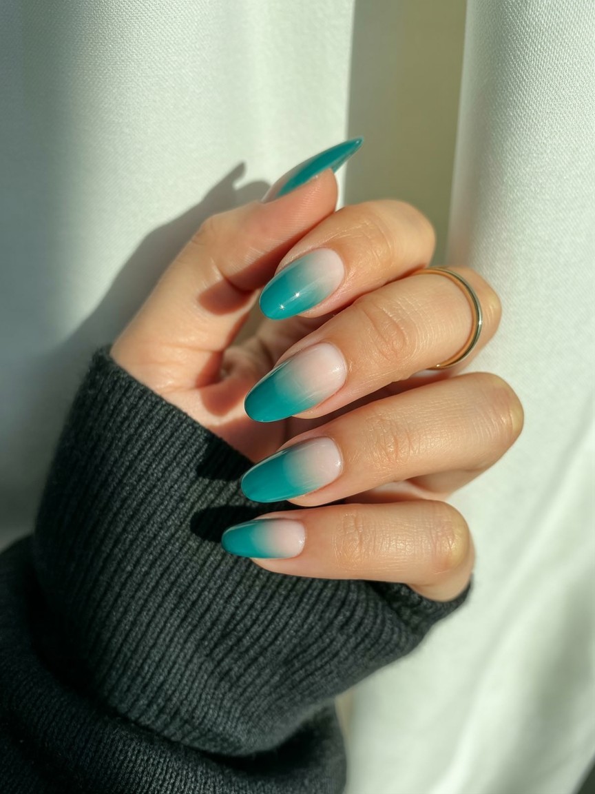 Side view of a hand with teal to clear ombre French tip nails, glossy finish, styled with a gold ring and dark knit sleeve on a neutral background.