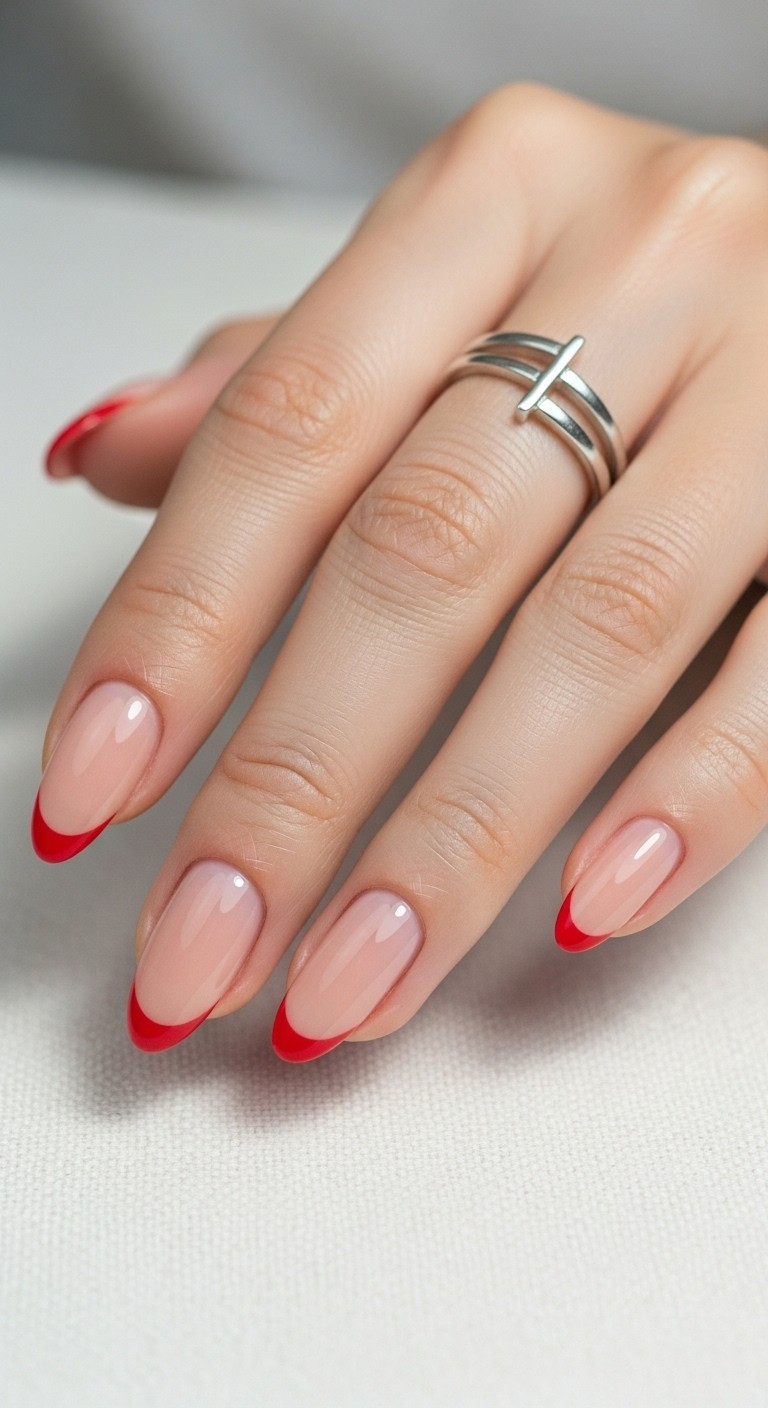 Hand with almond-shaped nude nails featuring thin, precise red French tips, styled with a minimalist silver ring.