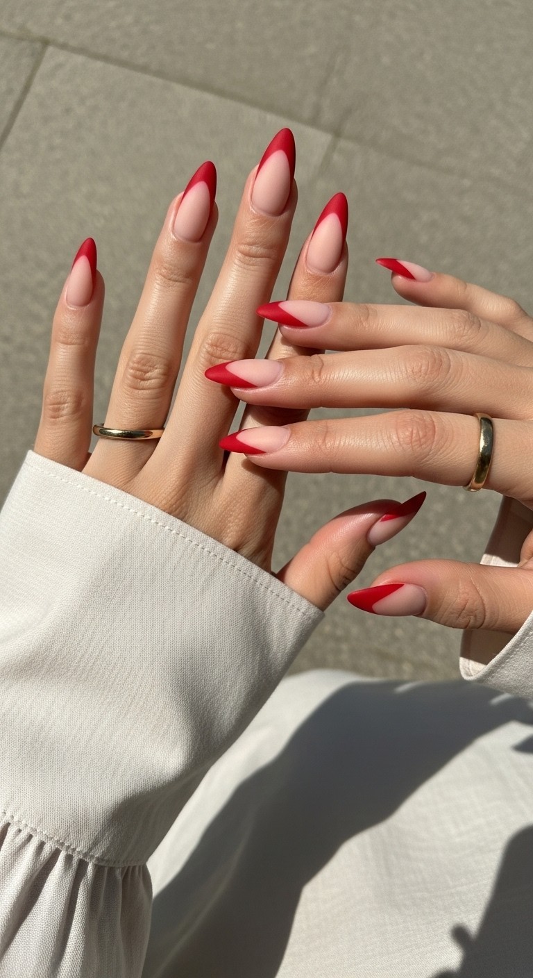 Almond-shaped nails with matte nude base and bold V-shaped red French tips, styled with minimal gold jewelry and soft neutral fabric sleeve.