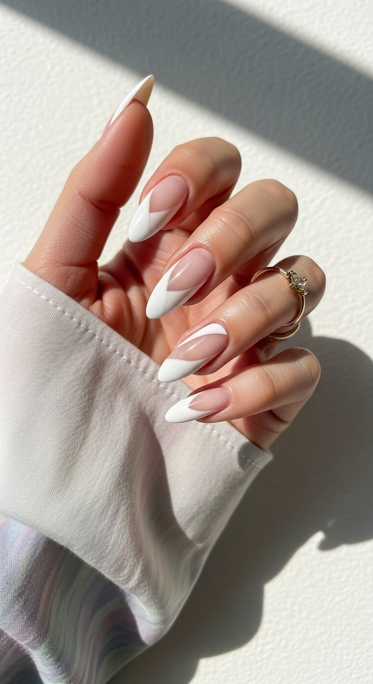 Hand with almond-shaped nails featuring asymmetrical white side tips on a nude base, accented with a delicate gold ring and framed by a pastel flowy sleeve, set against a soft off-white fabric background.