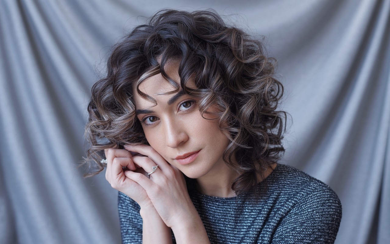 Curly dark brown hair with ash brown balayage highlights, styled with a silver ring and cool-toned sleeve against a soft gray fabric background.