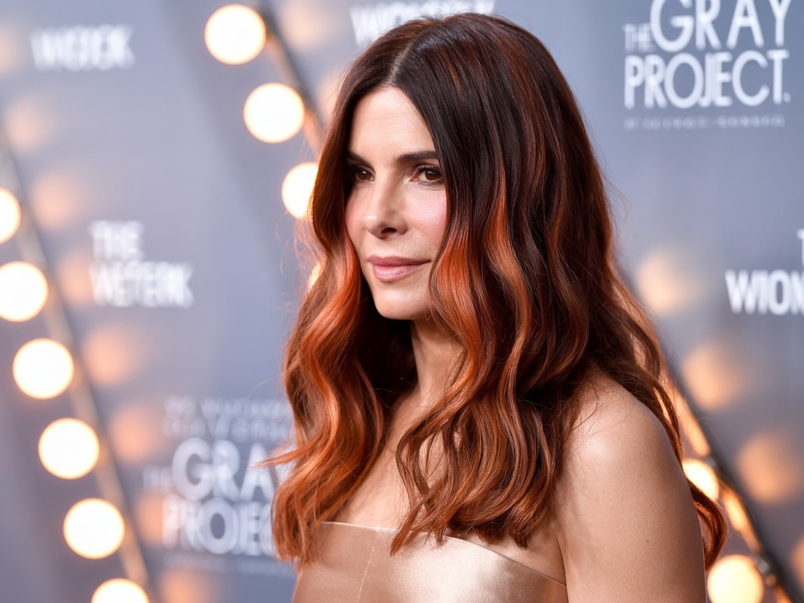 Sandra Bullock with seamless copper balayage on auburn hair, styled in soft, flowing waves with blended golden and amber highlights at a red carpet event.