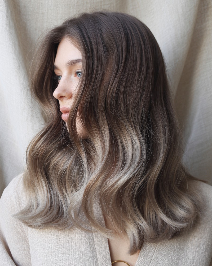 Soft ash brown balayage on long brunette hair with smoky beige ends, styled in polished waves to show a seamless color melt