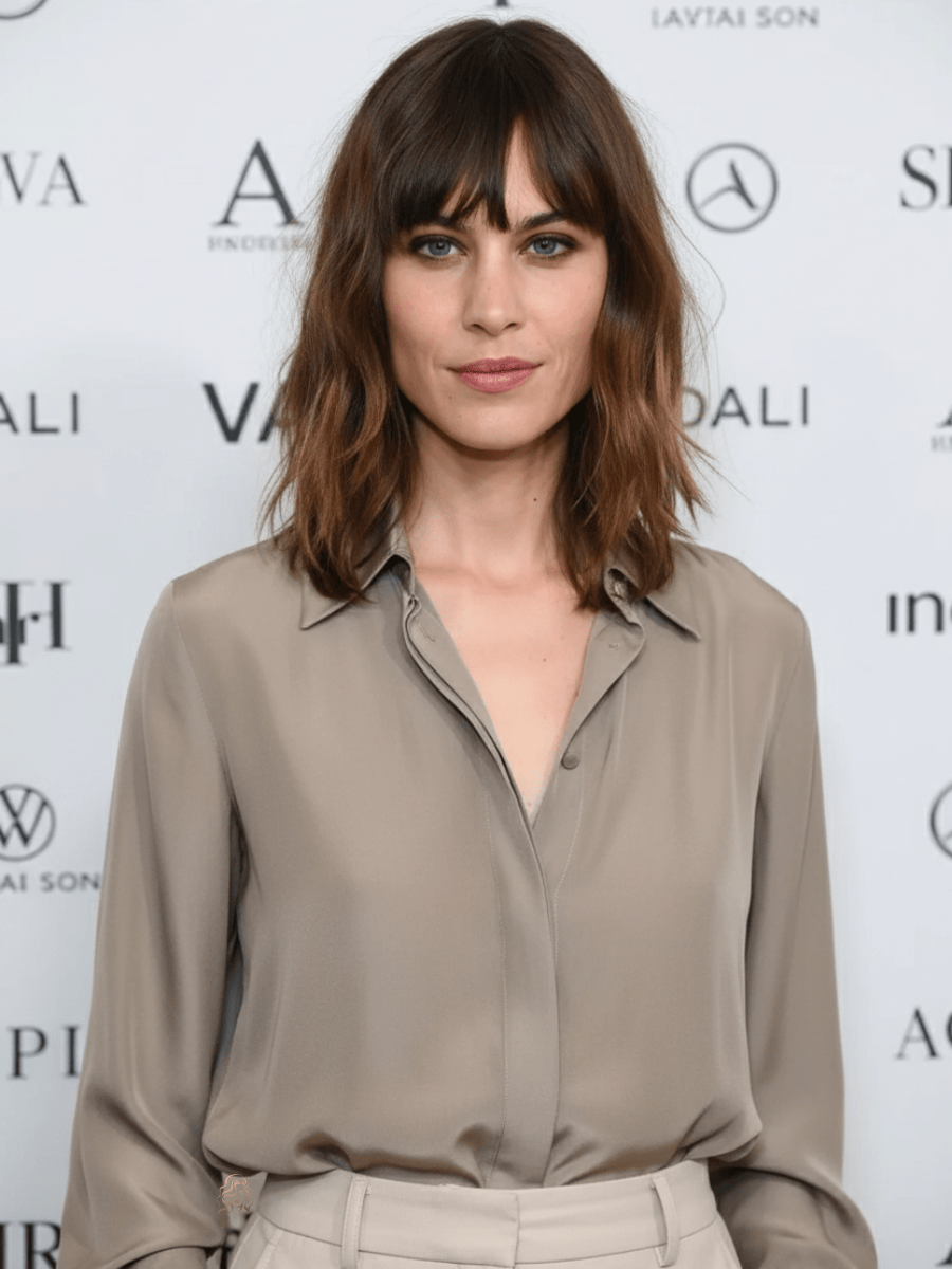 Alexa Chung standing at a fashion event backdrop, wearing a taupe silk blouse and tailored trousers, her shoulder-length modern shag haircut styled with natural waves and soft bangs framing her face.