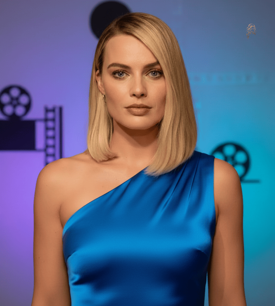 Margot Robbie wearing a cobalt blue satin one-shoulder gown with a sleek asymmetrical blonde lob, standing against a softly lit studio backdrop featuring film reel graphics.