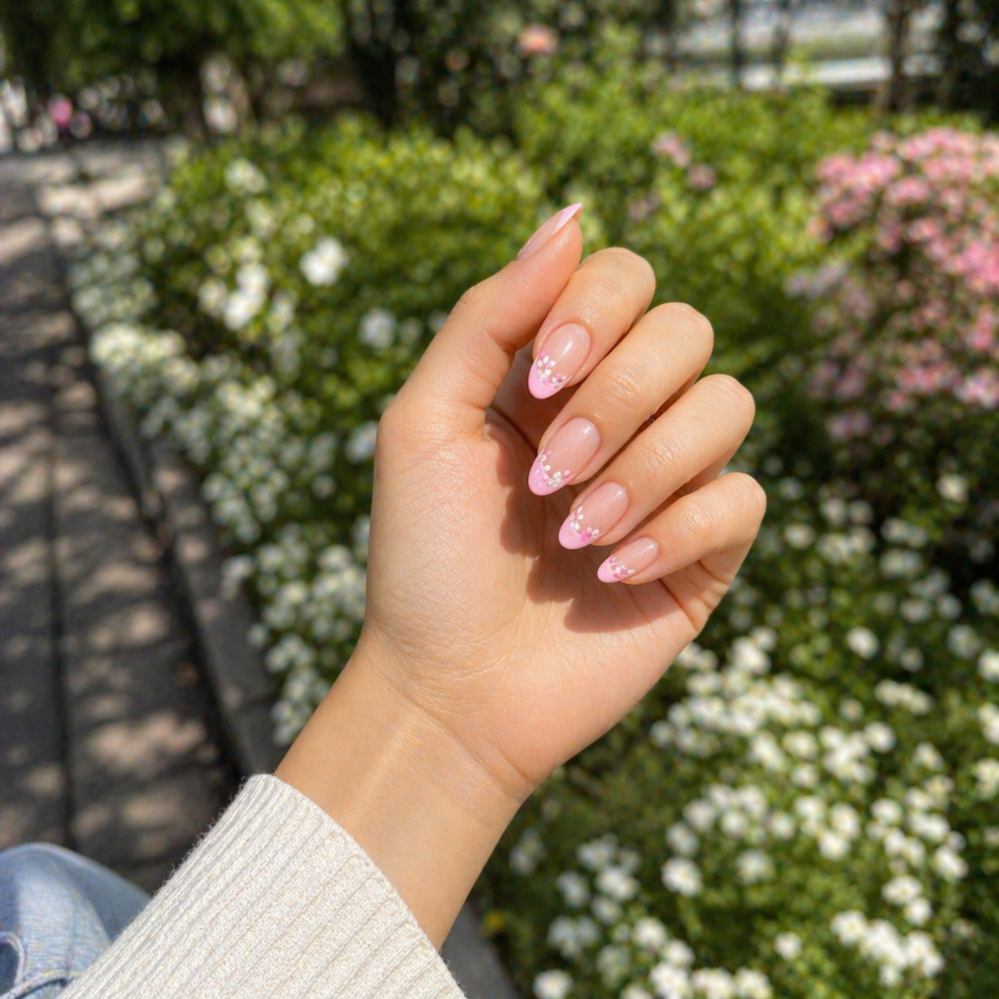 Almond nails with baby pink French tips and delicate floral accents in a soft outdoor spring setting with natural sunlight and a candid POV angle