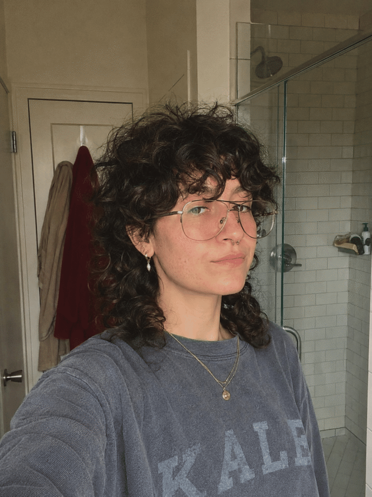 Day-three curly jellyfish haircut with natural lived-in texture, slightly elongated curls, soft crown frizz, and visible layered shape photographed in a bathroom mirror selfie with natural light.