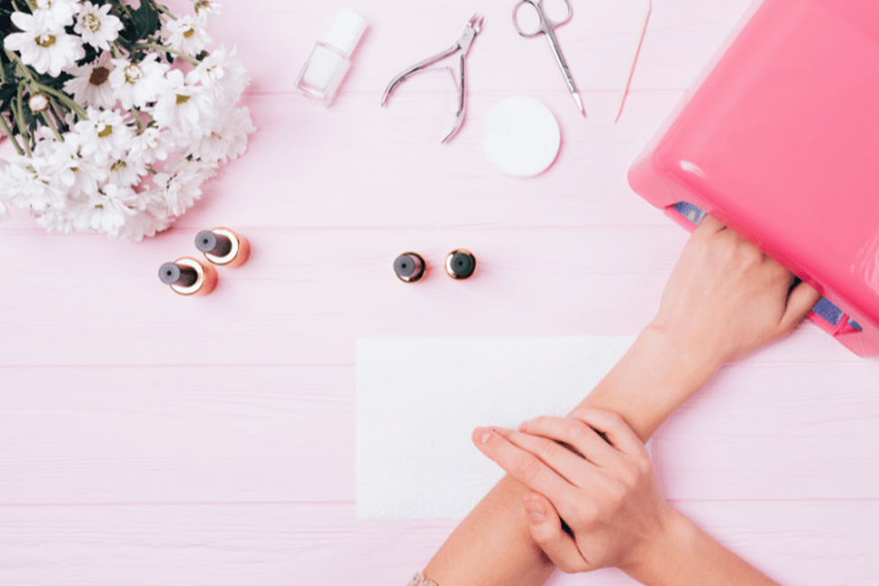 Hand curing gel nails under a professional UV nail lamp with manicure tools and nail polish on a salon workstation.